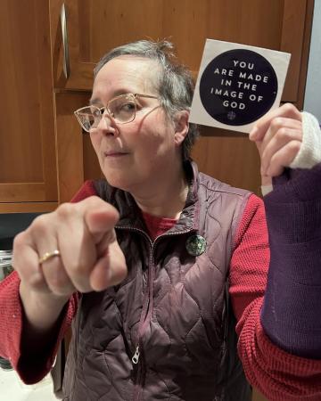 Patricia Bouteneff is holding a sticker that says, "You are made in the image of God" and is pointing at the viewer